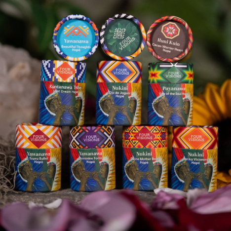 Stacked colorful Four Visions Hap&eacute; containers with patterned lids and labels, arranged among flower petals against a leafy green background.